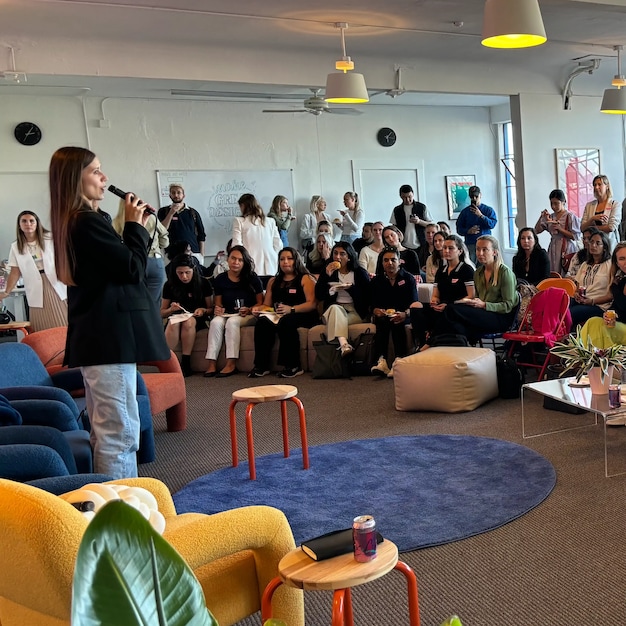 Speaker in black blazer presenting to a full audience in a modern office space with yellow chairs and blue rug